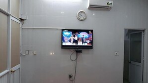 A room with a mounted TV displaying a show, an air conditioner, a clock, and a door. - Snooze Hotel (Hue)