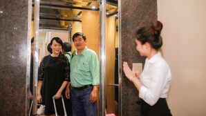 A group of people in an elevator with a woman in a white shirt and black skirt, a man in a green shirt and jeans, and a woman in a black dress with a white handbag. - Snooze Hotel (Hue)