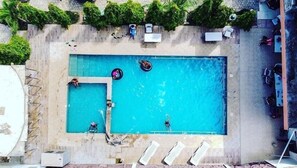 Vista aerea di una piscina rettangolare con una piccola zona adiacente, circondata da lettini e ombrelloni.