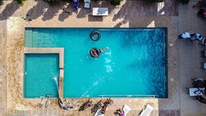 En vy från ovan av en rektangulär swimmingpool med ett mindre, intilliggande bubbelbad, omgivet av solstolar och parasoller.