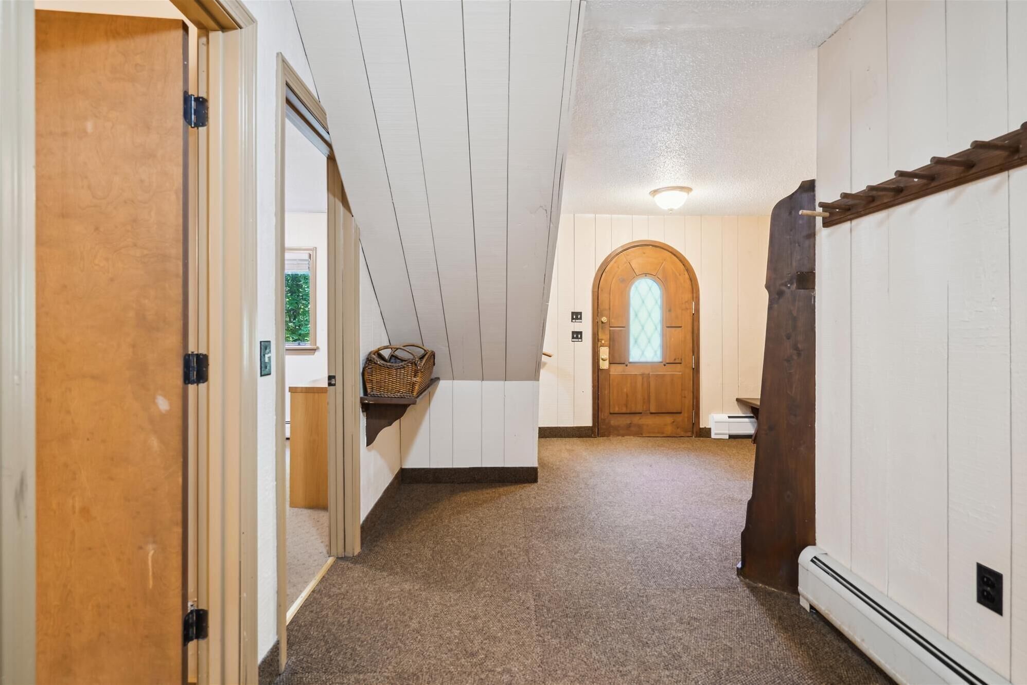 A hallway with a wooden door on the left, a white wall with a window, and a wooden door at the end.
