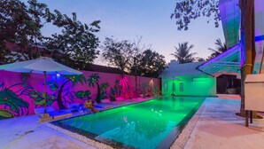 Une piscine ornée d’une fresque murale aux couleurs vives, entourée d’arbres et bordée d’un bâtiment dont l’entrée est illuminée de lumière verte.