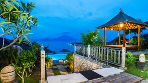 A gazebo with a stone pathway leading to a pool area, surrounded by tropical plants and overlooking a body of water with boats. - Jenggala Hill (Lembongan Island)