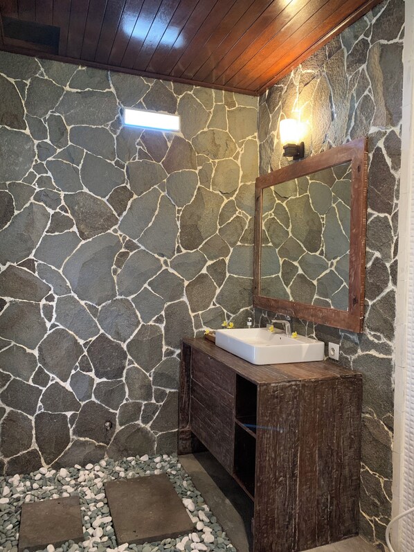A bathroom with a stone wall, wooden vanity, and a mirror. - Jenggala Hill (Lembongan Island)