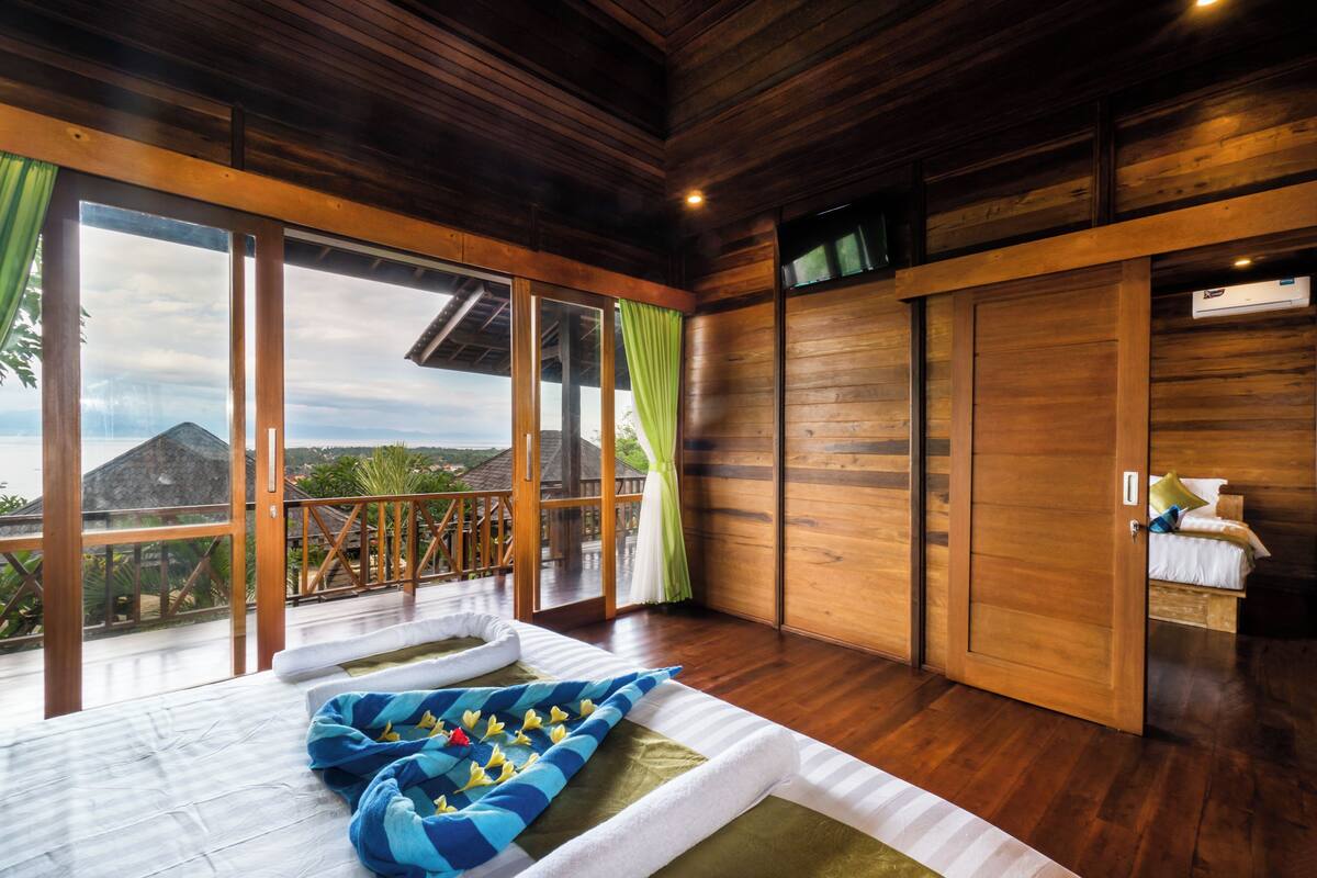 A room with a wooden floor, a bed with a blue and white striped blanket, a wooden wardrobe, and a large window with a view of the outdoors.