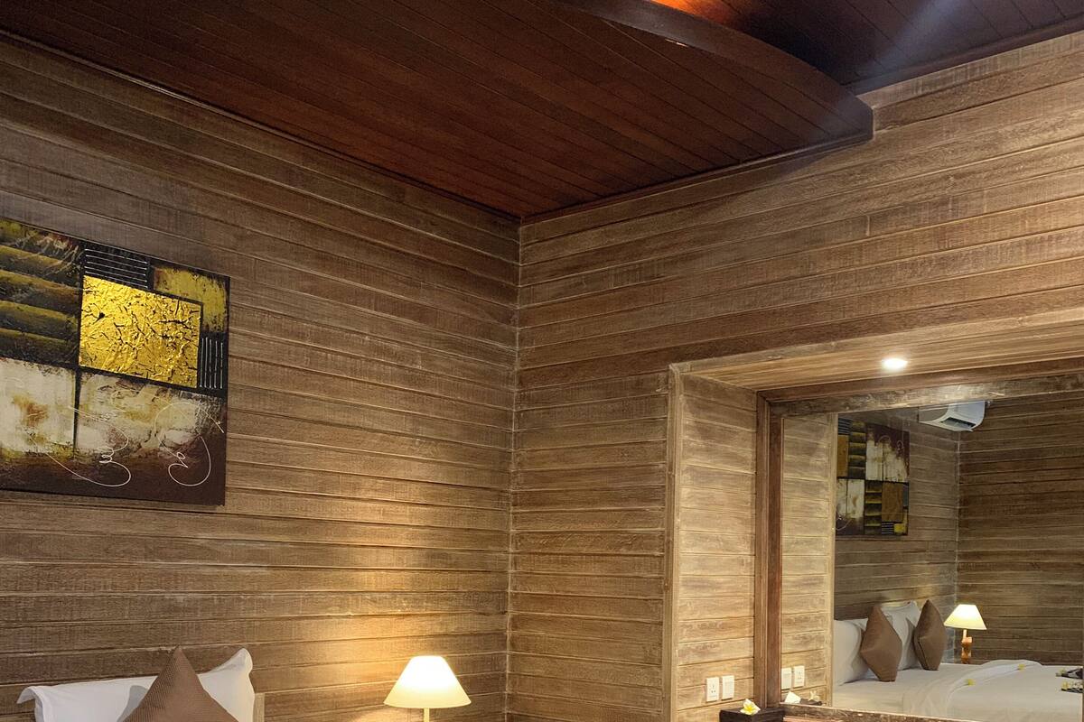 A hotel room with a wooden wall, a bed with white bedding, a bedside table with a lamp, and a mirror.