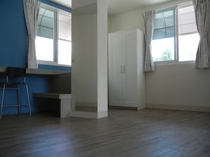 A room with a desk, chair, wardrobe, and two windows with curtains.