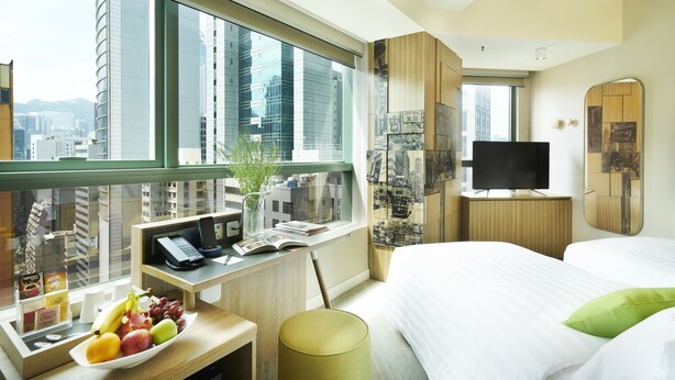 Una habitación de hotel con un ventanal grande, un escritorio con teléfono y un libro, una cama con ropa de cama blanca y un televisor sobre un atril de madera.