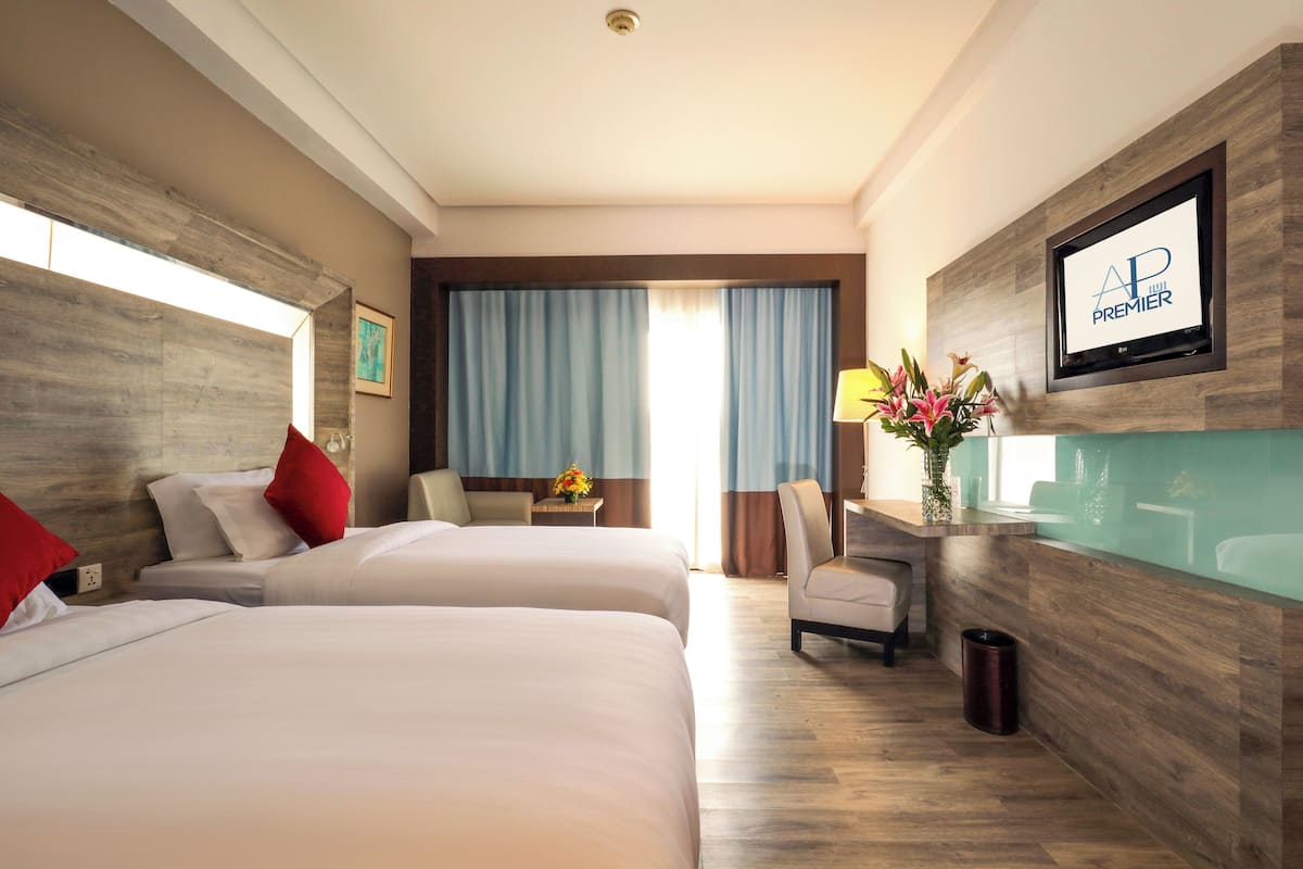 A hotel room with two beds, a desk with a lamp, a trash can, and a large window with curtains.