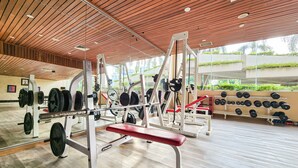 A gym with weightlifting equipment, a red bench, and a mirrored wall reflecting the outdoor greenery. - AP Premier Batam (Batam)