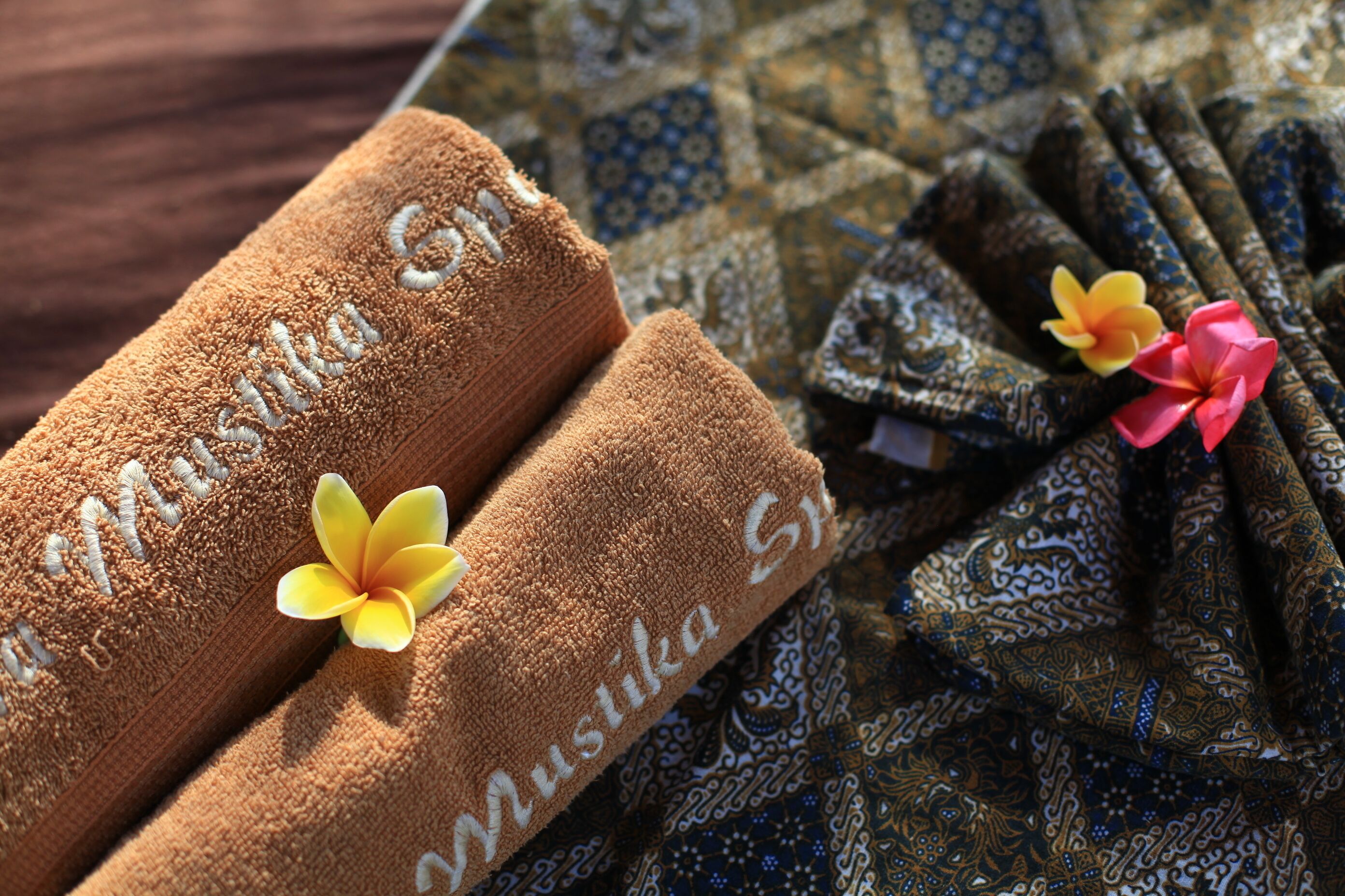 Two towels with 'Mustika Spa' embroidered on them, placed on a patterned fabric with a floral decoration.