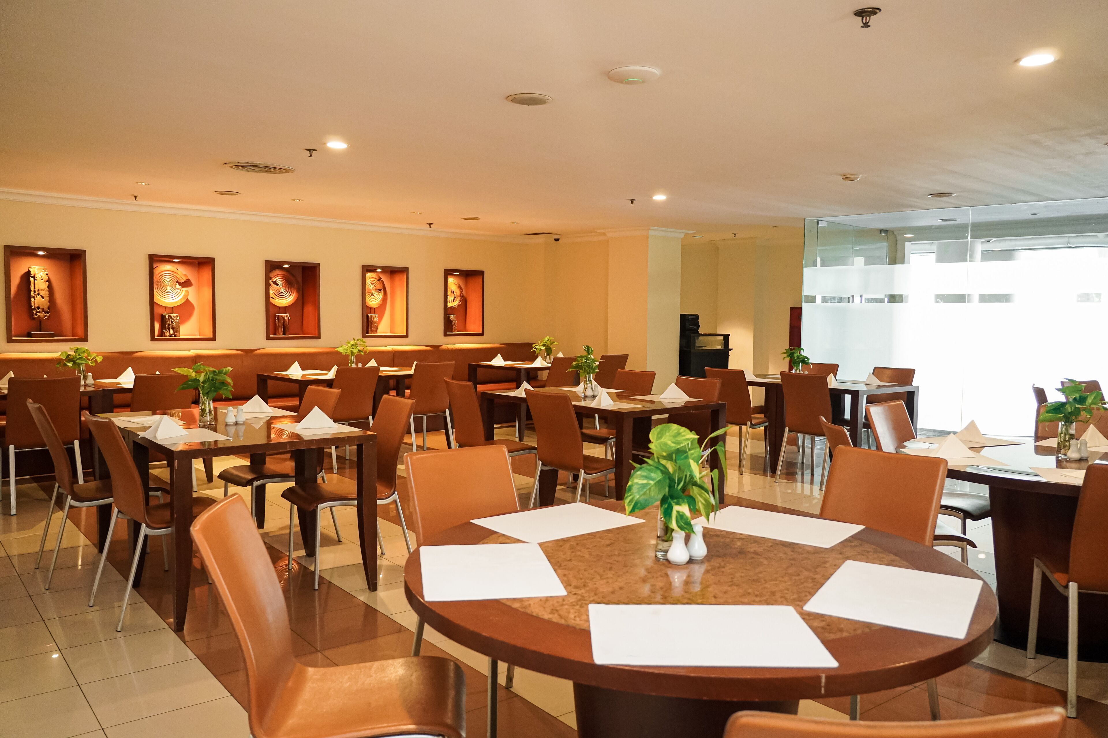 A hotel dining area with multiple tables and chairs, framed artwork on the walls, and potted plants.