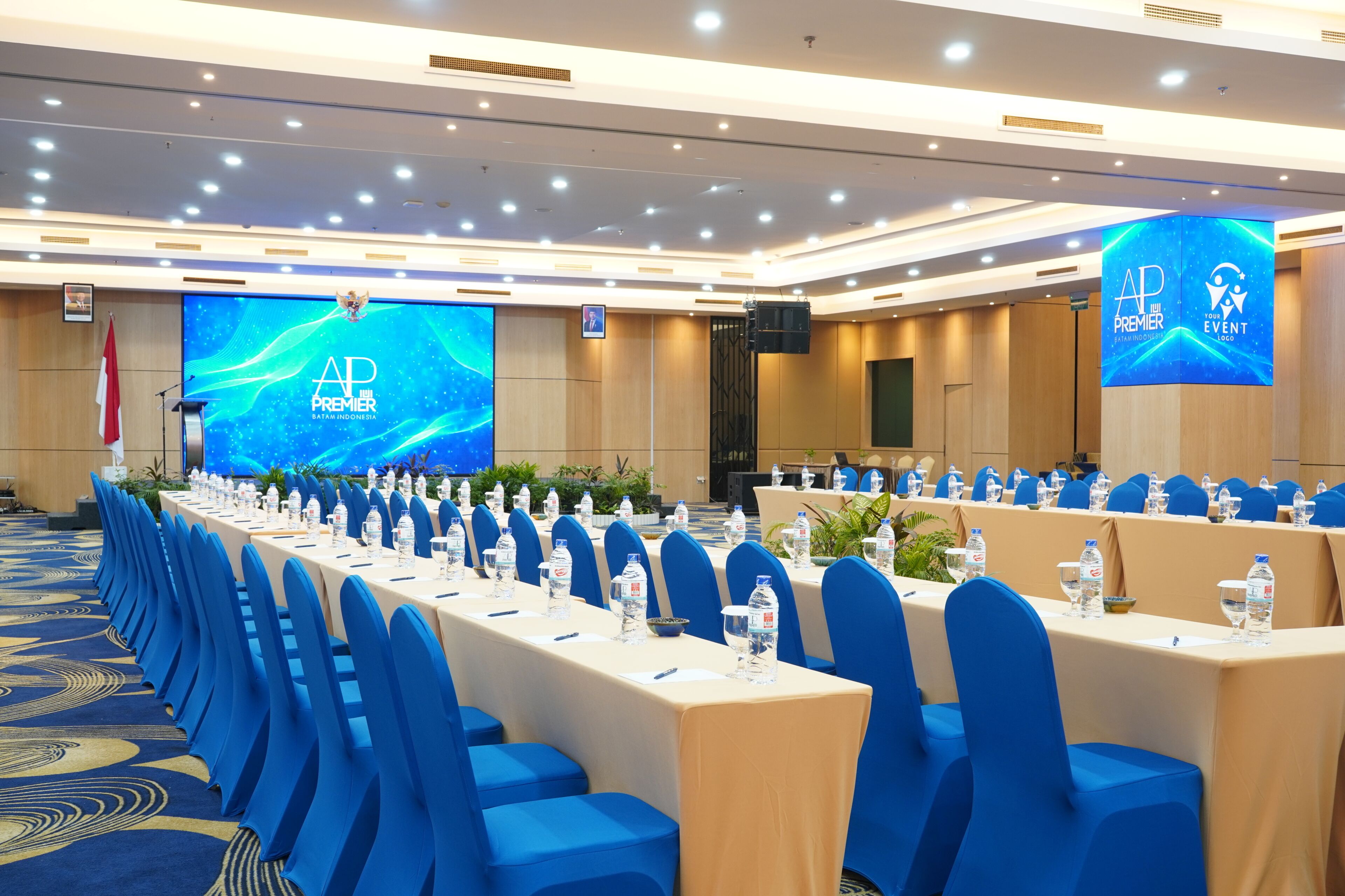 Une salle de conférence avec une longue table préparée pour une réunion, des chaises bleues et un grand écran affichant le texte « AP PREMIER BANTAM INDONESIA ».