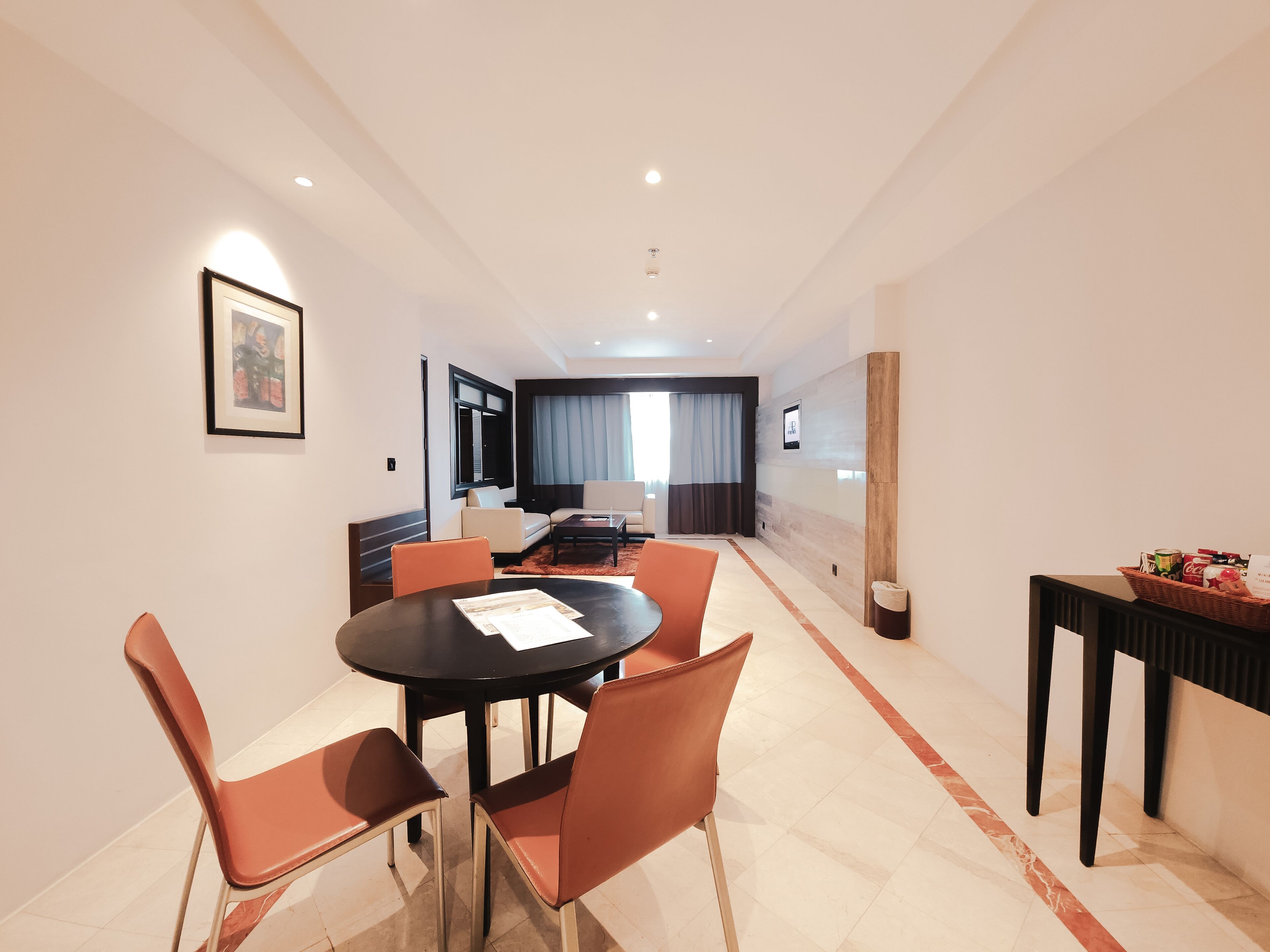 A modern hotel room with a dining area, a sofa, a large window with curtains, and a reception desk with a basket of items.
