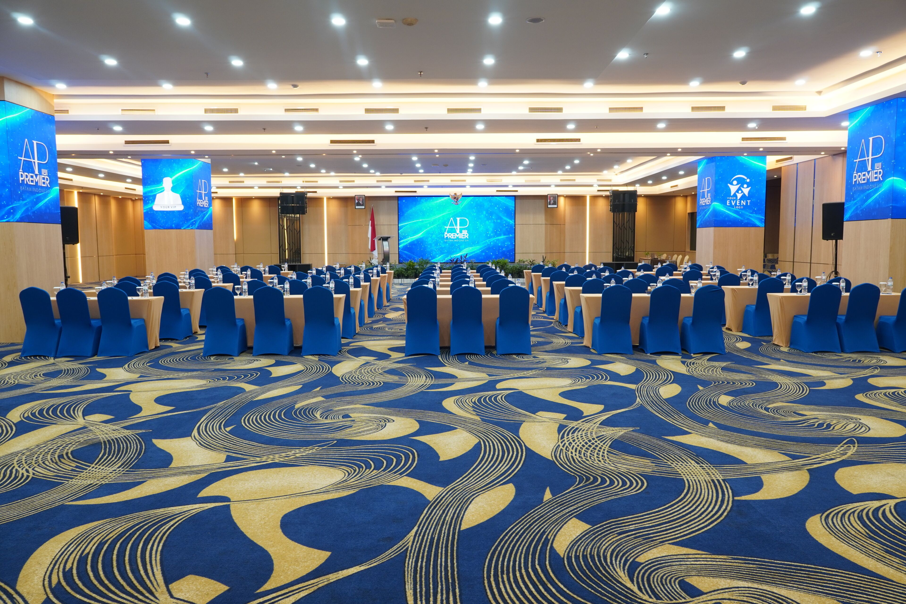Une salle de conférence avec des rangées de chaises bleues, un grand écran à l’avant et un tapis à motifs.