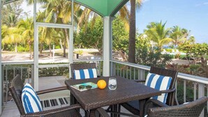 A covered outdoor seating area with wicker furniture and a table, overlooking a tropical landscape with palm trees and a pool.