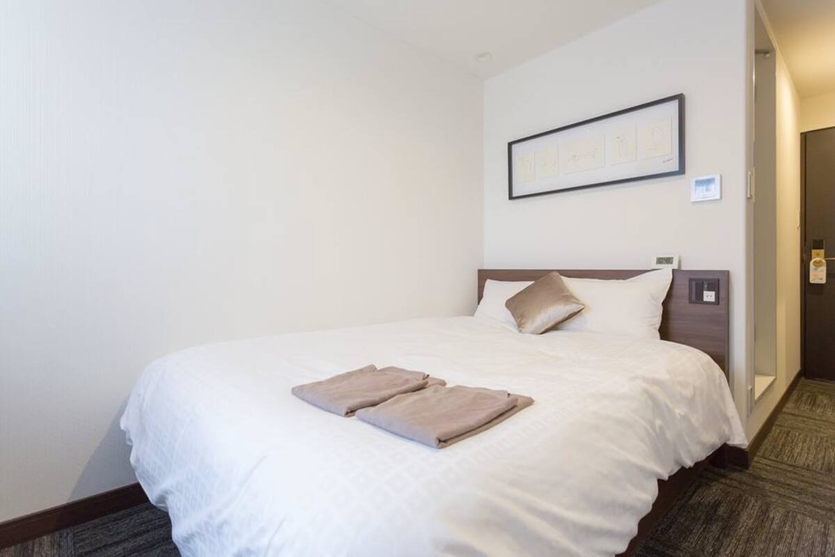A neatly made bed with a white comforter and two beige towels, a framed picture on the wall, and a carpeted floor.
