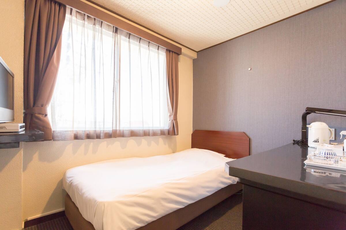 A hotel room with a single bed, a desk with a lamp, and a window with curtains.
