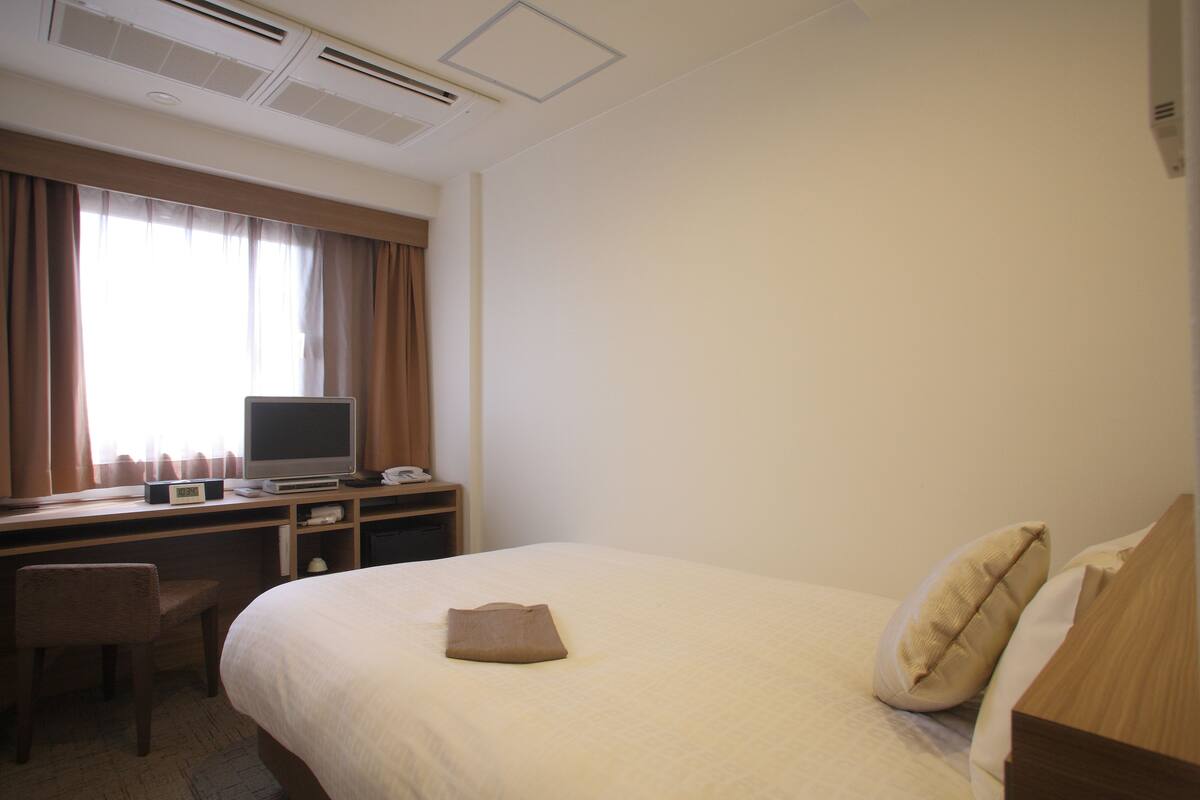 A hotel room with a bed, a desk with a computer, a chair, and a window with curtains.