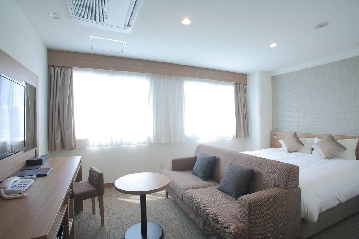 A hotel room with a bed, a sofa, a desk with a phone and a laptop, and a TV.