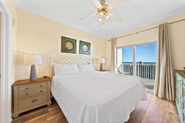 A bedroom with a large bed, two bedside tables with lamps, a ceiling fan, and a balcony with curtains.