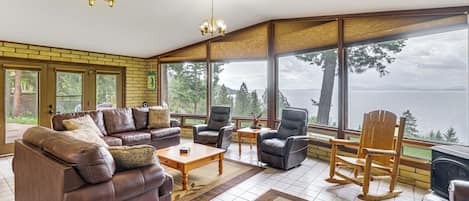 A spacious living room with a large window overlooking a body of water, furnished with leather sofas, chairs, and a wooden rocking chair.