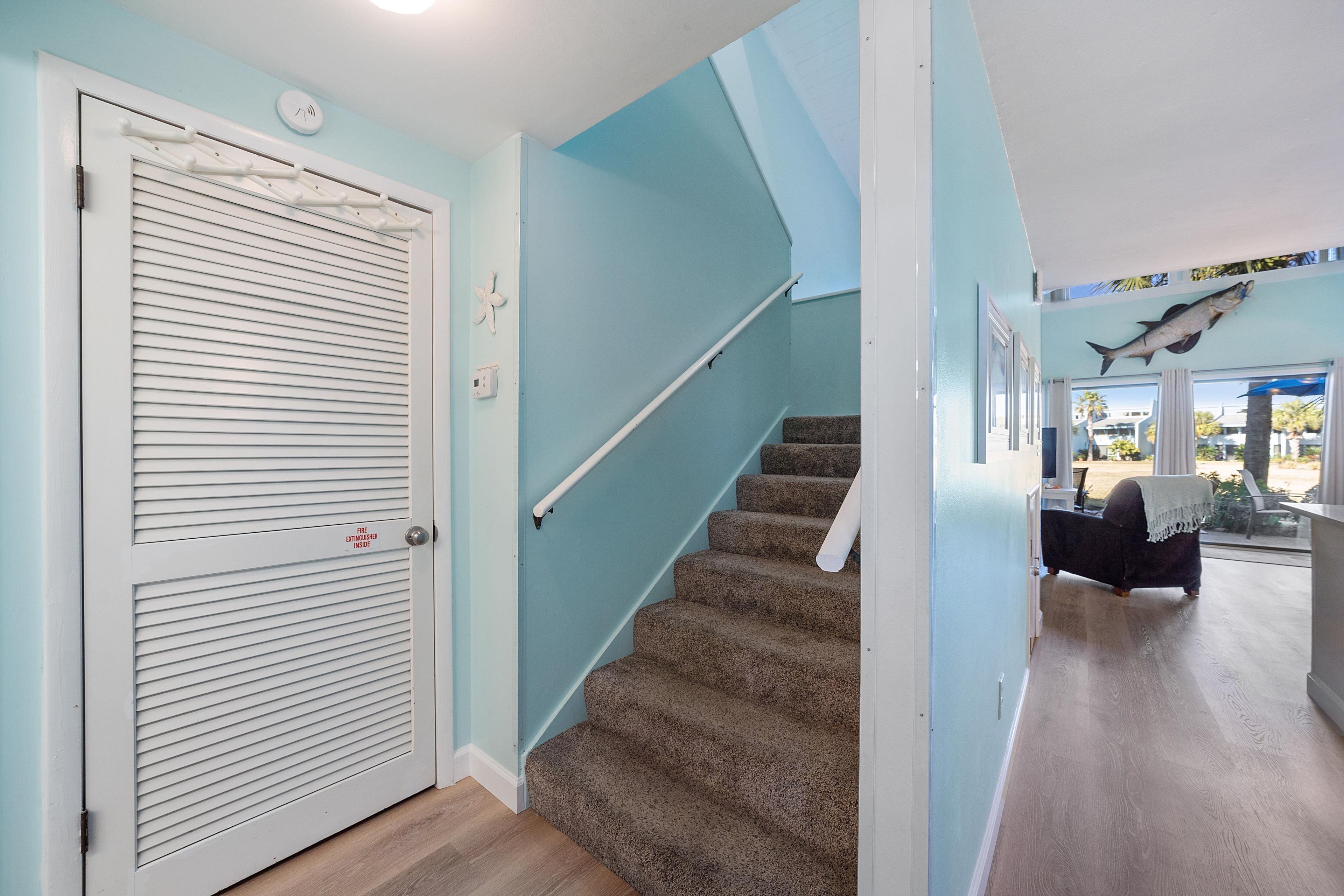 A staircase with a handrail, a closed door with a fire alarm, and a view of a room with a bed and a window.