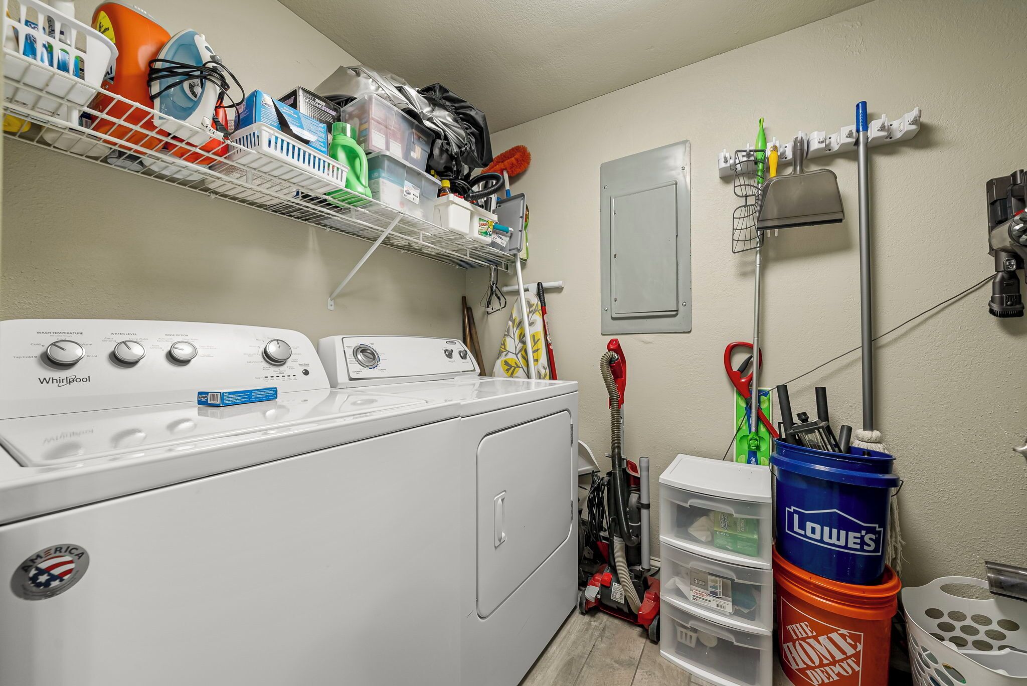 Une buanderie équipée d’un lave-linge et d’un sèche-linge Whirlpool, d’étagères contenant des produits de nettoyage et de bacs de rangement.