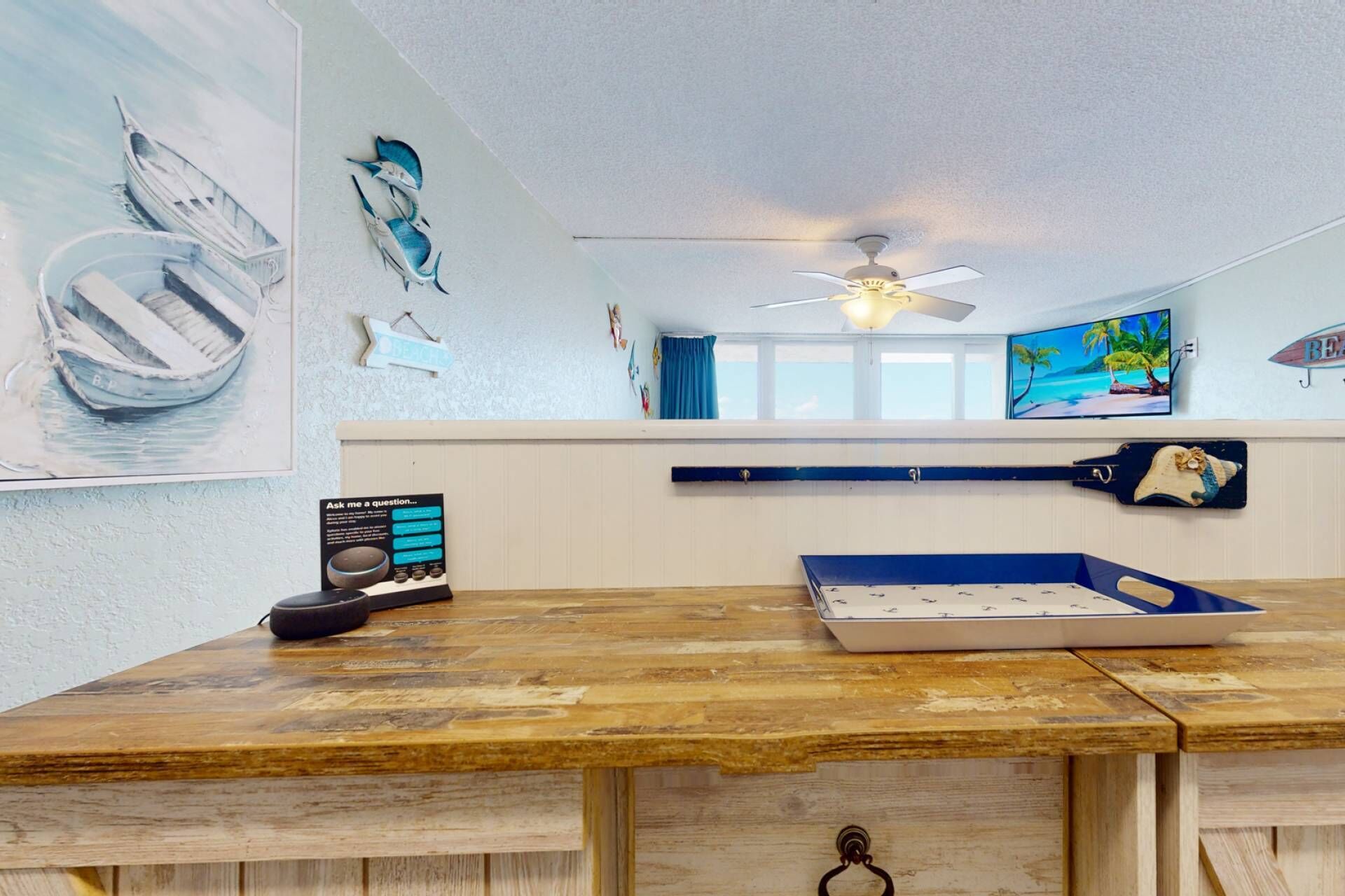 A room with a wooden desk, a ceiling fan, a TV displaying a beach scene, and a wall with nautical-themed artwork.