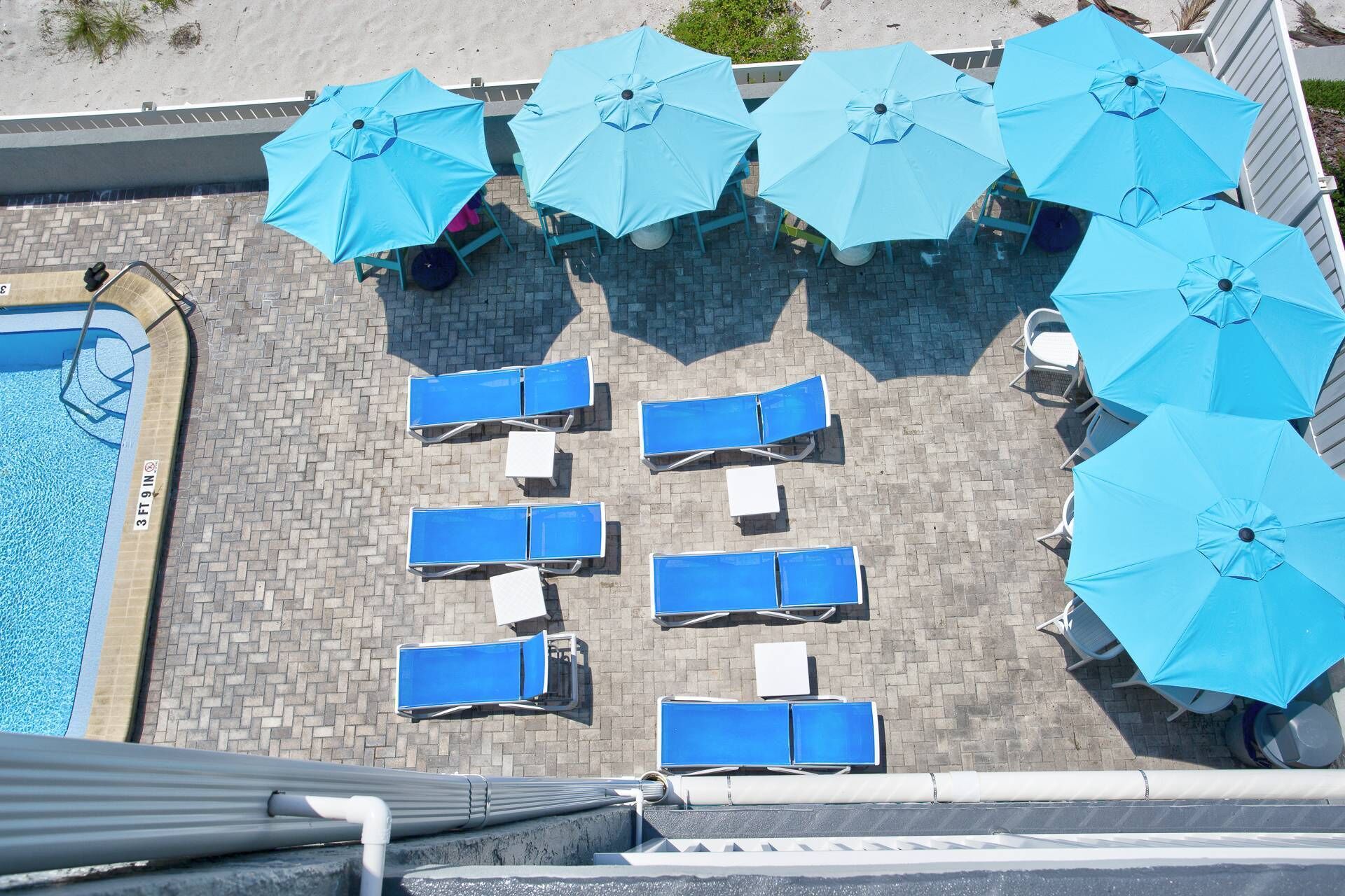 An aerial view of a poolside area with multiple blue umbrellas, lounge chairs, and a swimming pool.