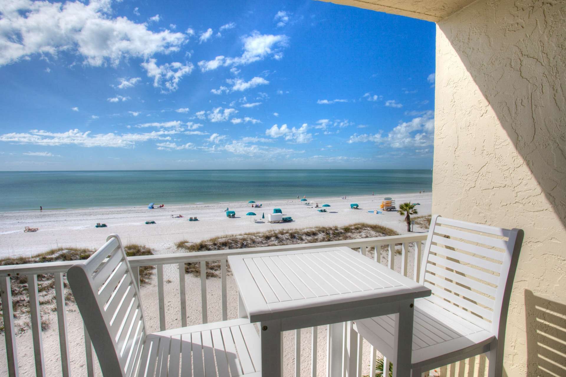 Een balkon met witte meubels met uitzicht op een strand met diverse parasols en strandstoelen.