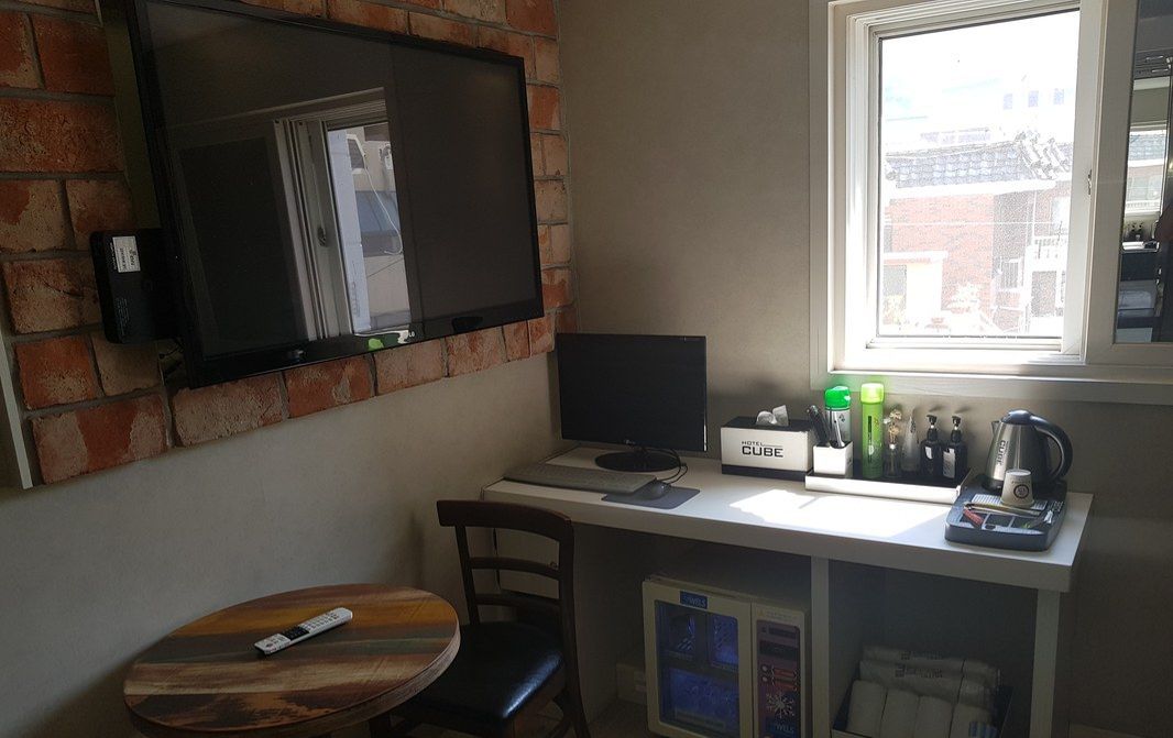 A small, compact room with a brick wall, a flat-screen TV mounted above a desk, a window with a view of buildings, a small round table, and a chair.