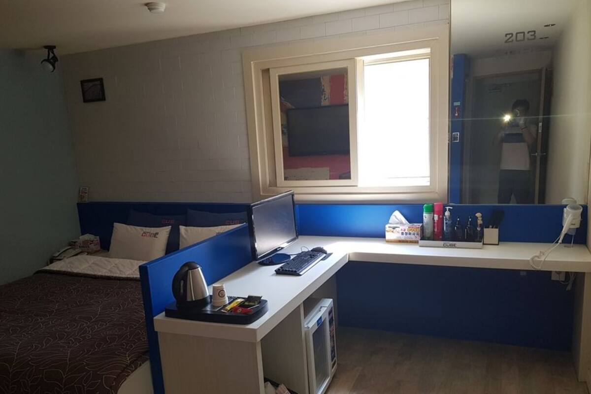 Una habitación con cama, escritorio con computadora portátil, ventana y espejo.