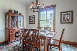 A dining room with a wooden table set for six, surrounded by chairs, a chandelier, and a large window with a view. - Lodges 1140 | Shuttle | Shared Hot Tub (Mammoth Lakes)