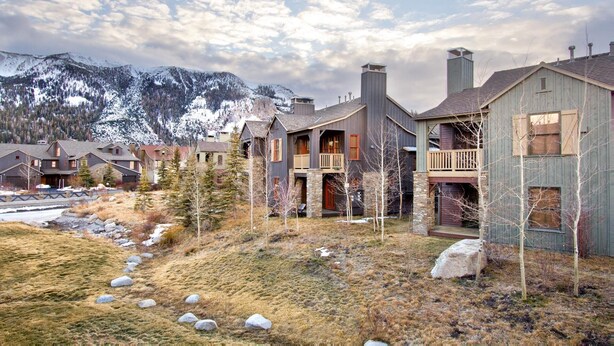 A row of rustic cabins with stone foundations, wooden exteriors, and balconies, set against a backdrop of snow-capped mountains and a clear sky. - Lodges 1140 | Shuttle | Shared Hot Tub (Mammoth Lakes)