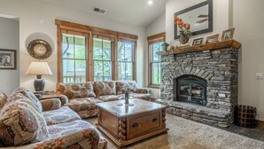 A living room with a stone fireplace, patterned sofas, a coffee table, and large windows. - Lodges 1140 | Shuttle | Shared Hot Tub (Mammoth Lakes)