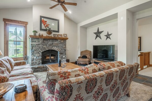 A living room with a stone fireplace, a television, and patterned sofas. - Lodges 1140 | Shuttle | Shared Hot Tub (Mammoth Lakes)