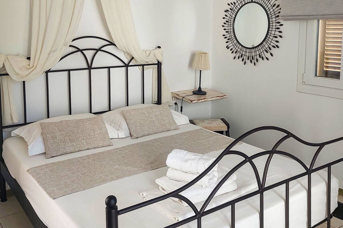 A neatly made bed with a headboard, a bedside table with a lamp, a wall-mounted air conditioner, and a decorative mirror.