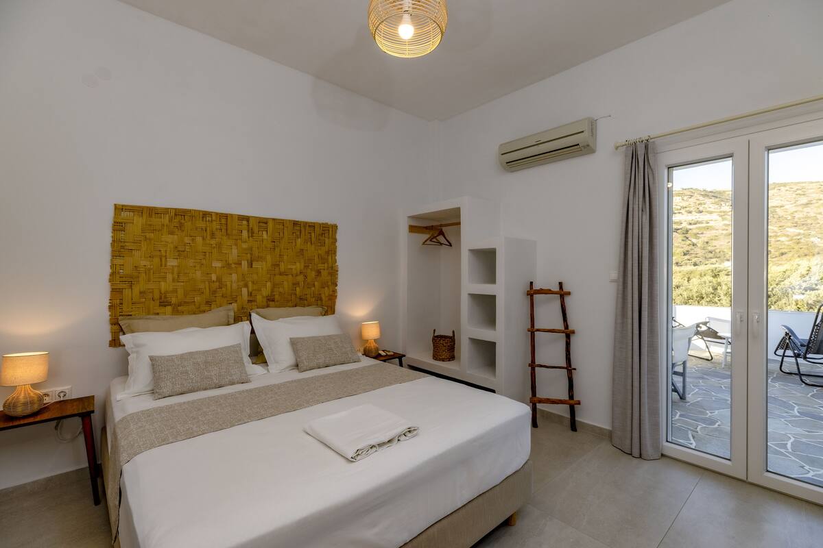 A neatly arranged bedroom with a bed, bedside tables, a hanging basket, and a view through a sliding glass door to an outdoor area.