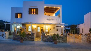A two-story building with a sign reading "Trini Pension" and "Seni Apartments". It features a stone facade, multiple windows with shutters, and a covered outdoor seating area. - Irini Pension (Sifnos)