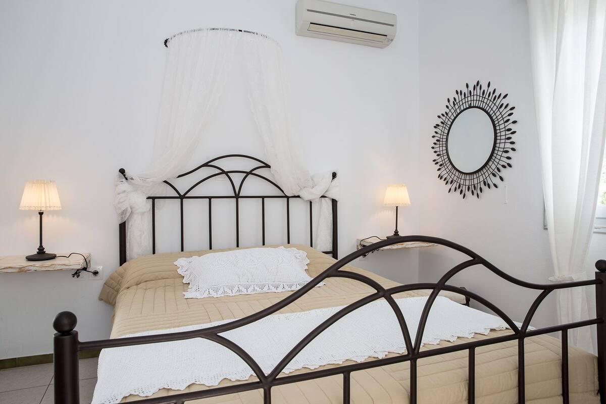 A neatly arranged bedroom with a bed, bedside lamps, a wall-mounted air conditioner, and a decorative mirror.