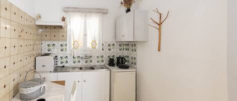 A compact kitchen with white cabinets, a small refrigerator, a sink, and a window with curtains.