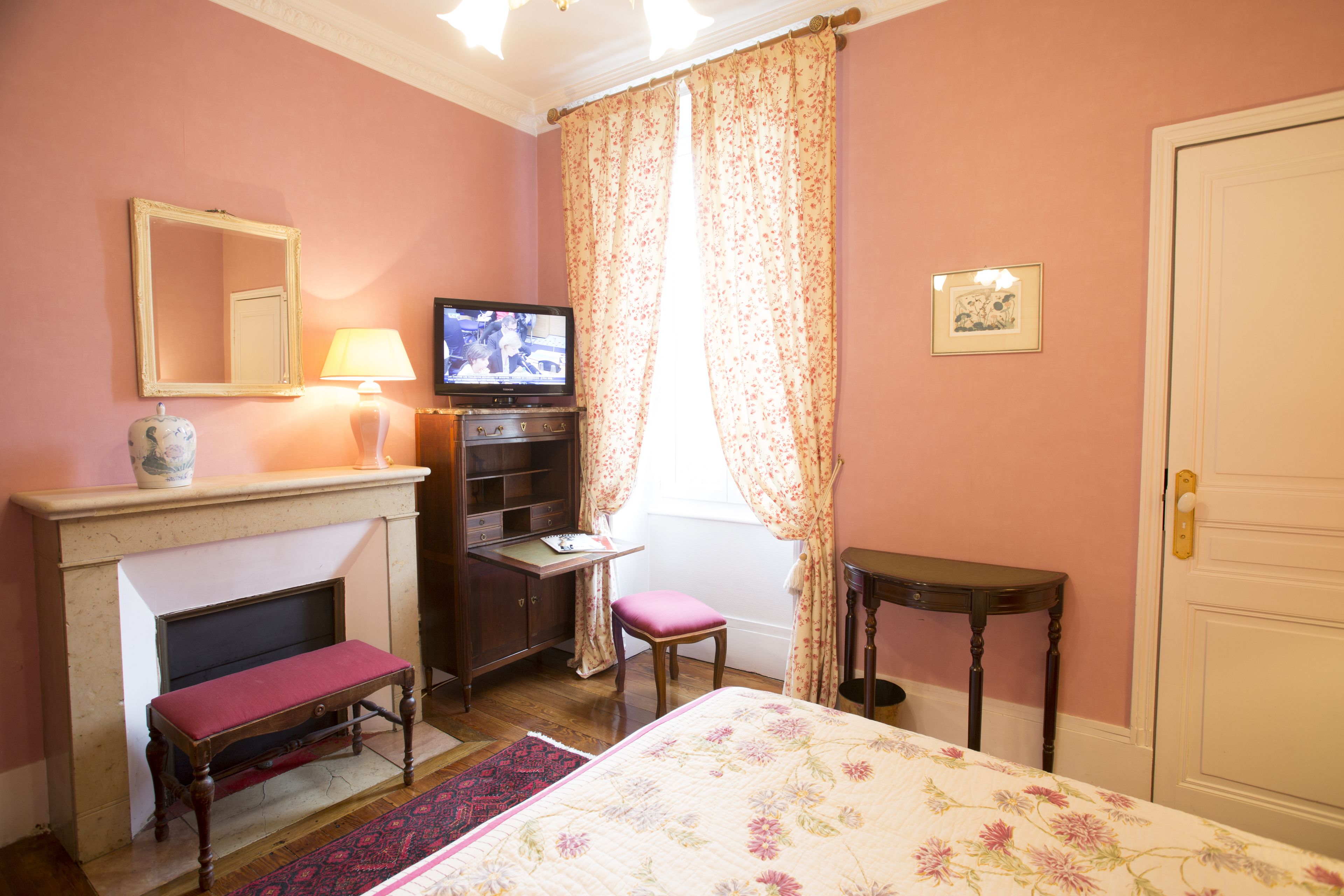 A bedroom with a bed, a television, a desk, a chair, a small table, a mirror, a fireplace, and a door.