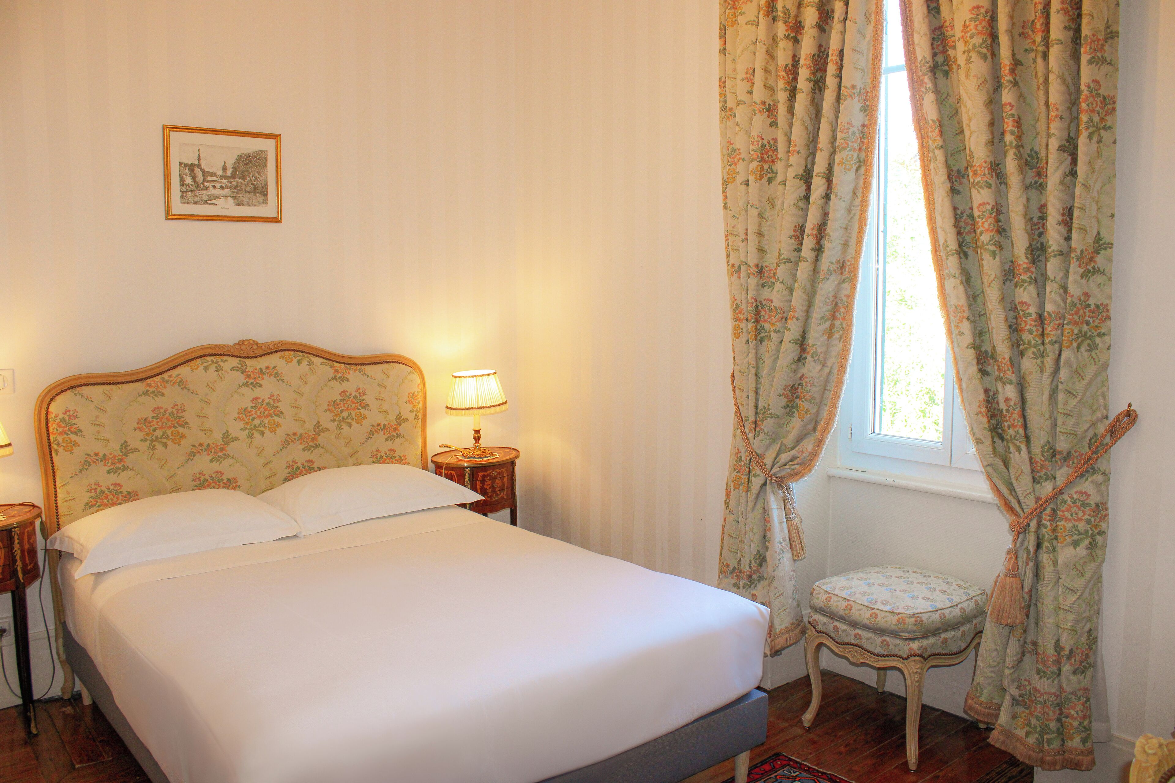 Una cama bien hecha con cabecera floral, una lámpara de noche y una pequeña silla junto a la ventana con cortinas estampadas.