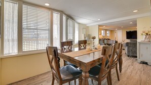 Un comedor con mesa y sillas de madera, ventanas grandes con persianas, una cocina al fondo y un televisor sobre un mueble.