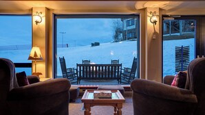 A cozy seating area with wicker chairs and a wooden table, overlooking a snowy outdoor scene with a building and a chairlift. - CX313 | Ski In/Out | Spacious Floorplan | Winter Shuttle | Great Amenities (Steamboat Springs)