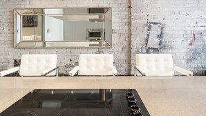 Une cuisine dotée d’une grande plaque de cuisson à induction, de trois chaises blanches à revêtement, et d’un grand miroir accroché à un mur en briques.