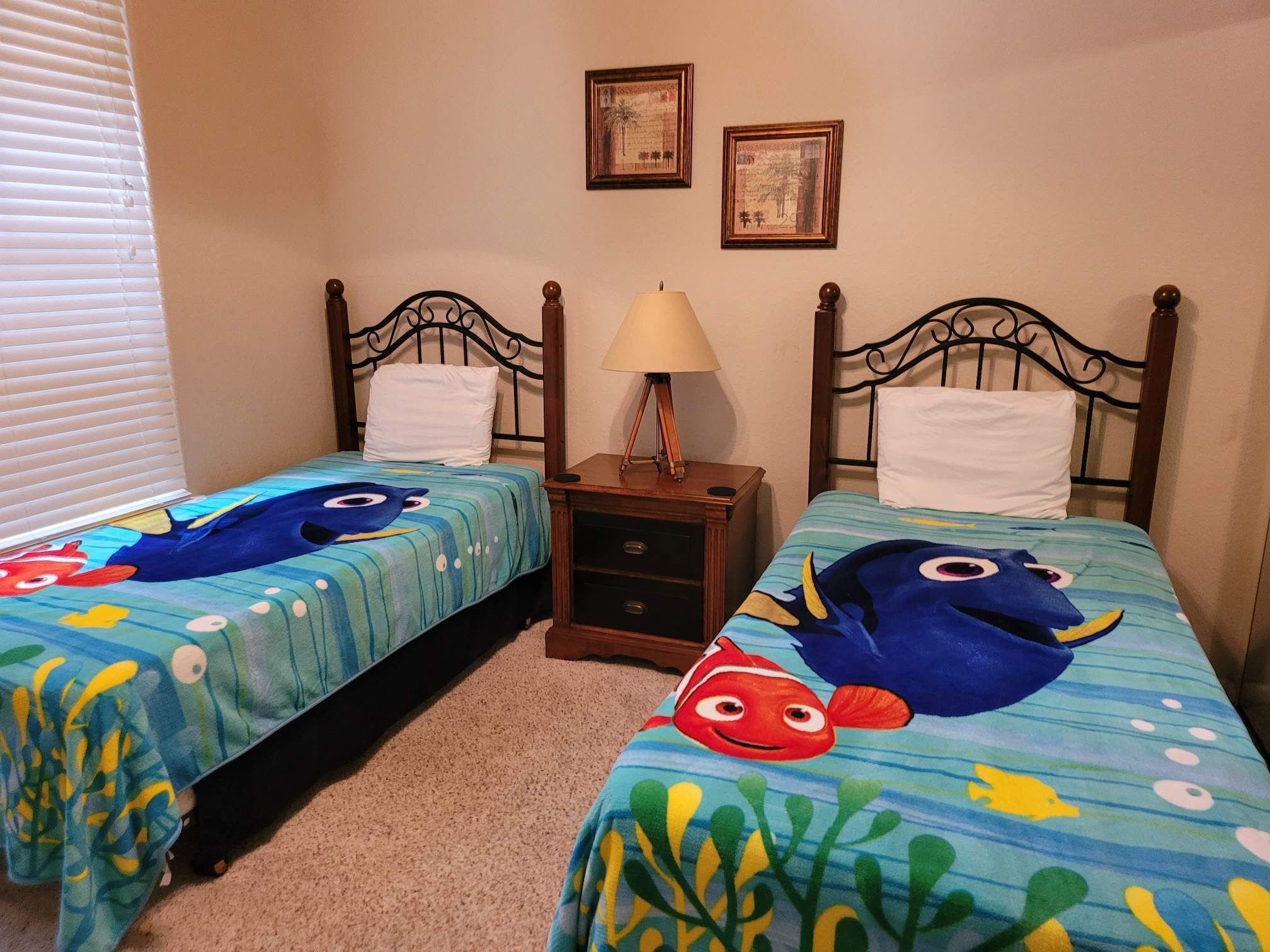 Une chambre avec deux lits, chacun recouvert d’une couvre-lit aux motifs de poissons bleus et jaunes, une table de chevet avec une lampe, et deux tableaux encadrés accrochés au mur.