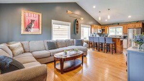 A modern living room with a sectional sofa, a wooden coffee table, and a kitchen area with wooden cabinets and a stainless steel refrigerator.