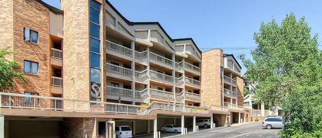 A multi-story building with a brick facade, multiple balconies, and a parking area with several cars.